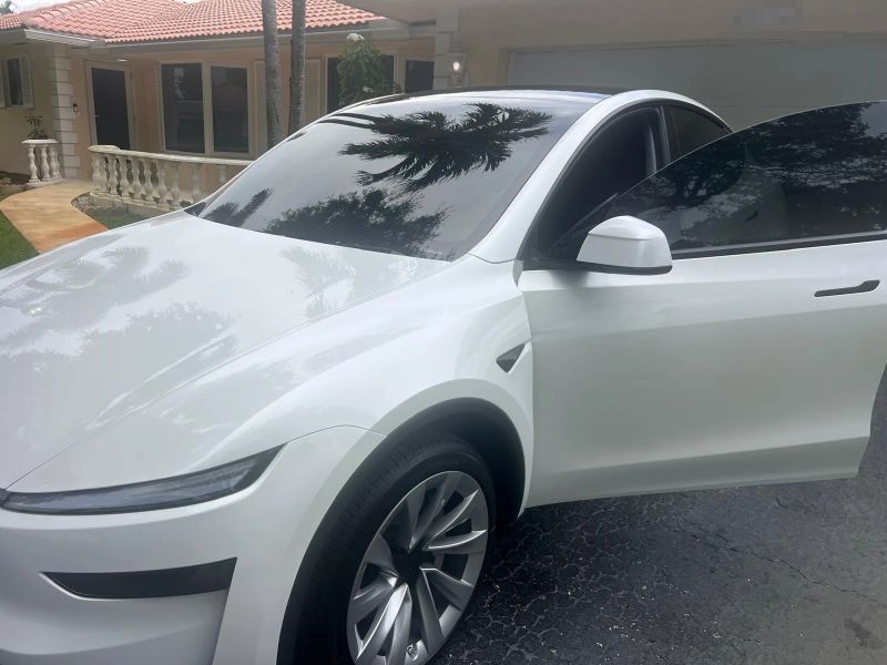 White new-style Tesla Model Y with dark tinted windshield and side glass reflecting palm trees